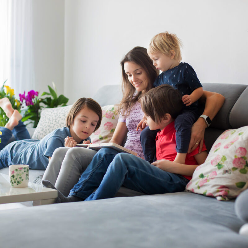 Young woman, mother with three kids, reading a book at home, hugging and laughing, family happiness