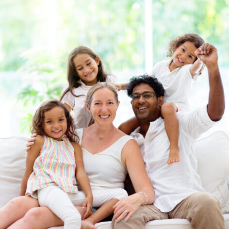 Happy family with kids at home. Parents and children sitting on white couch in sunny living room. Mother, father, son and daughter play and laugh. Young beautiful interracial couple in new house.